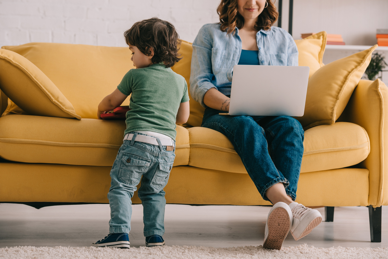 kid standing near sofa while mother working with laptop